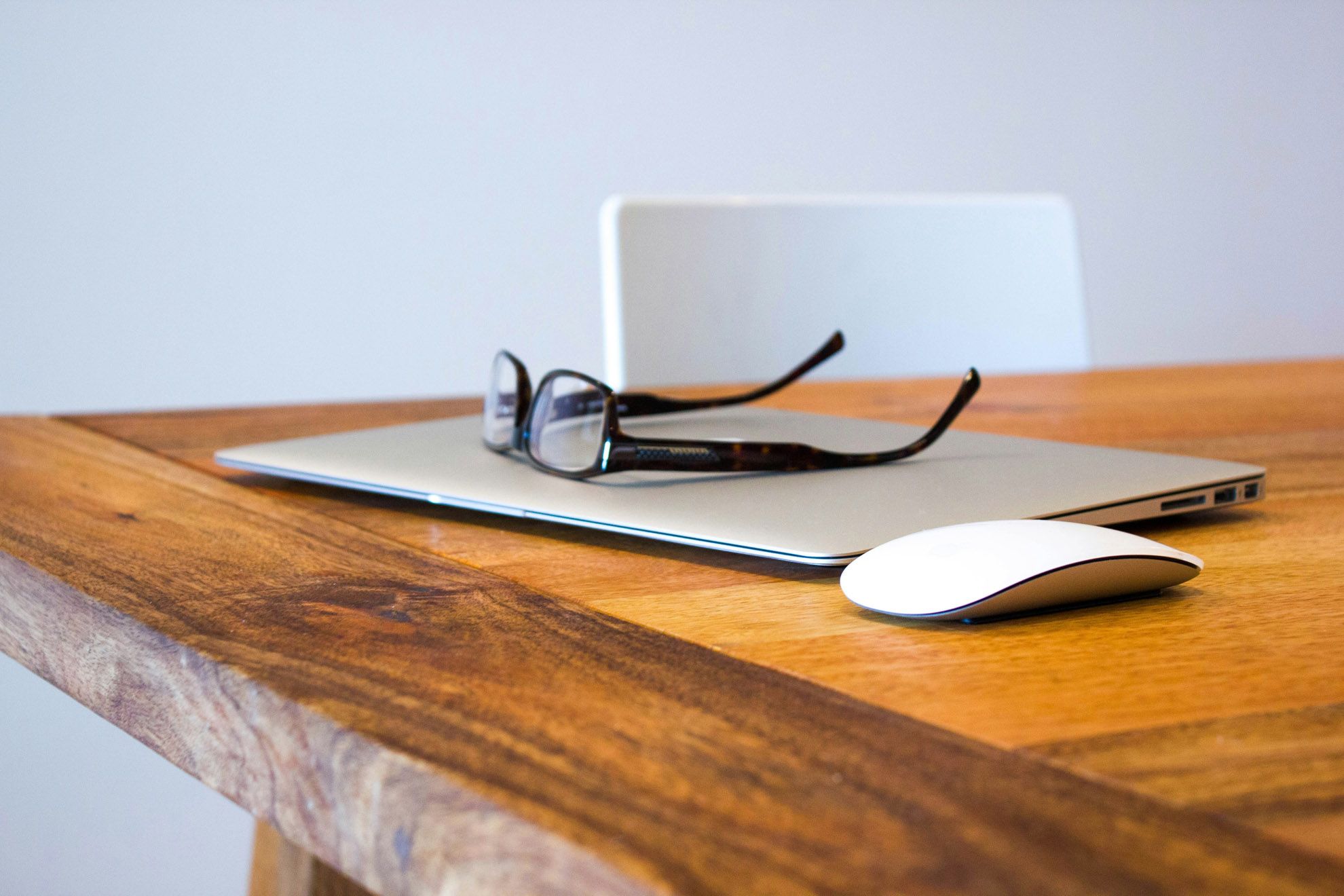 Picture of a closed silver laptop on a wooden desk. There are reading glasses placed on the laptop.